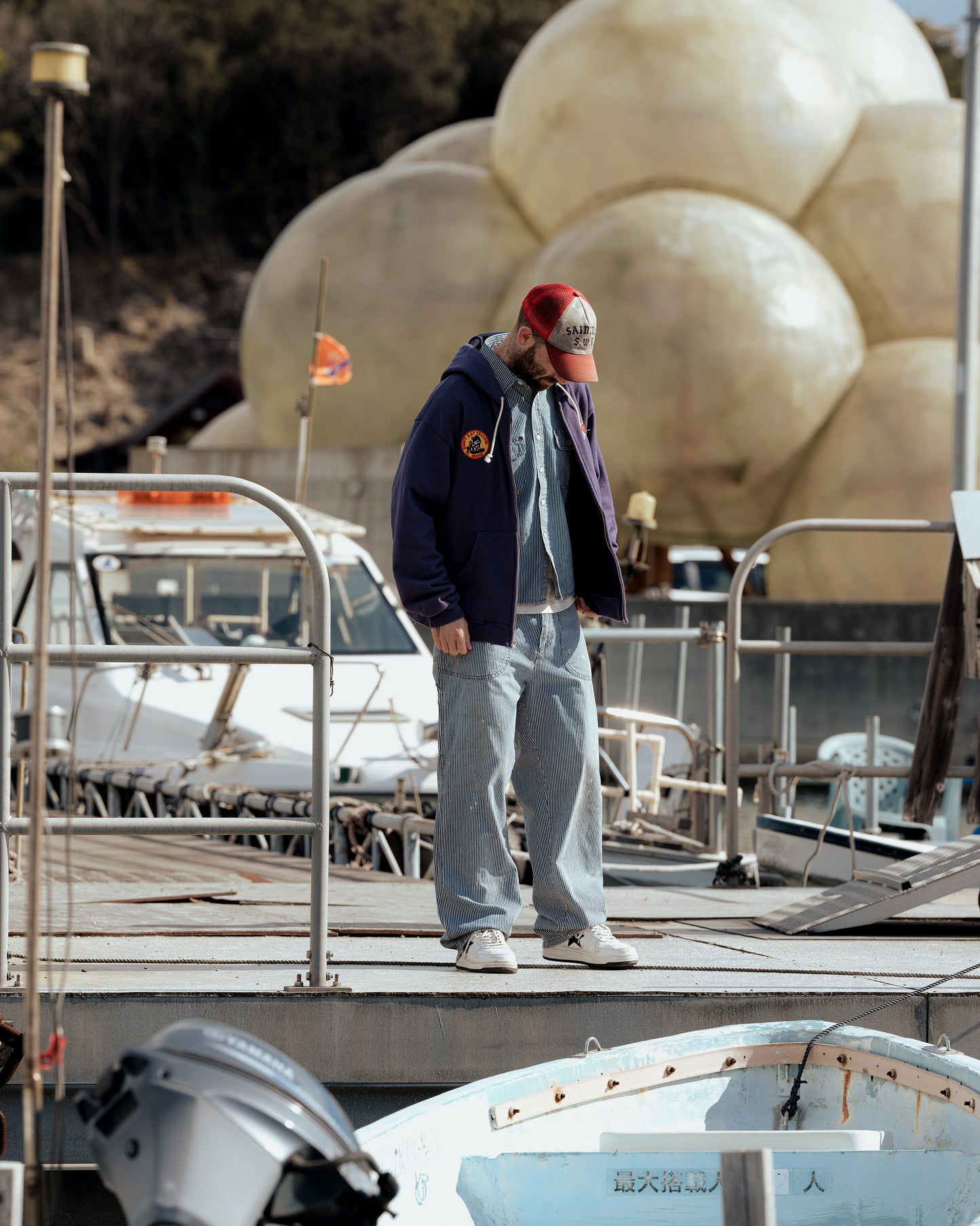 SWJP Zip Hoodie - Sean Wotherspoon Designs 公式。ネイビーフーディーを着た男性、港の桟橋でリラックスした雰囲気。