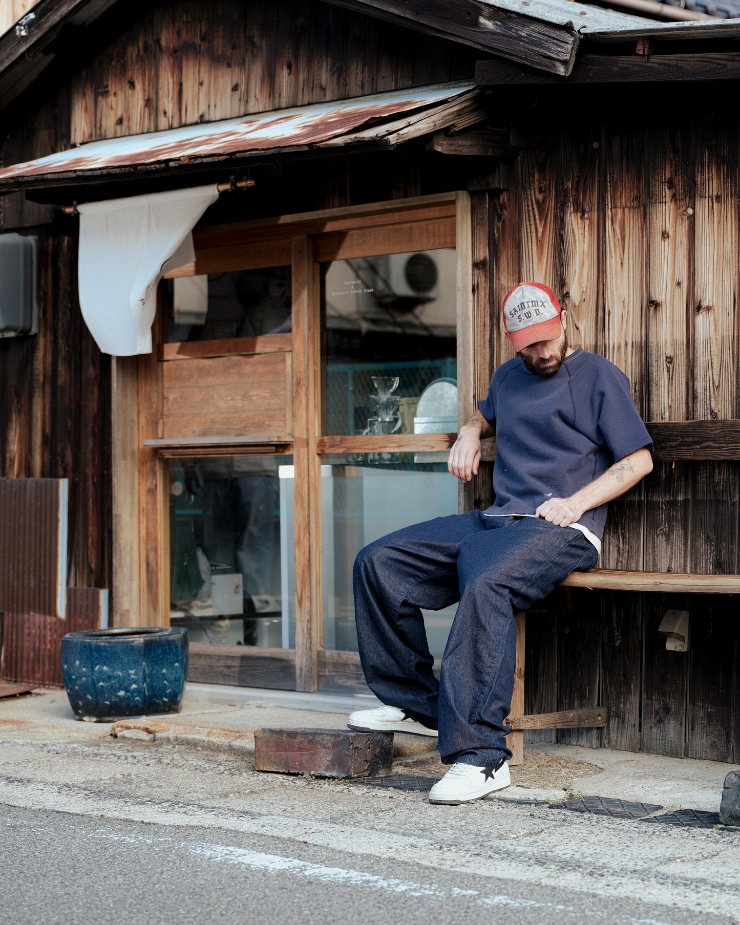 SWJPのS/S Sweatshirtを着た男性が木造の建物前で座る。ネイビーのショートスリーブスウェットがラフな雰囲気を演出。